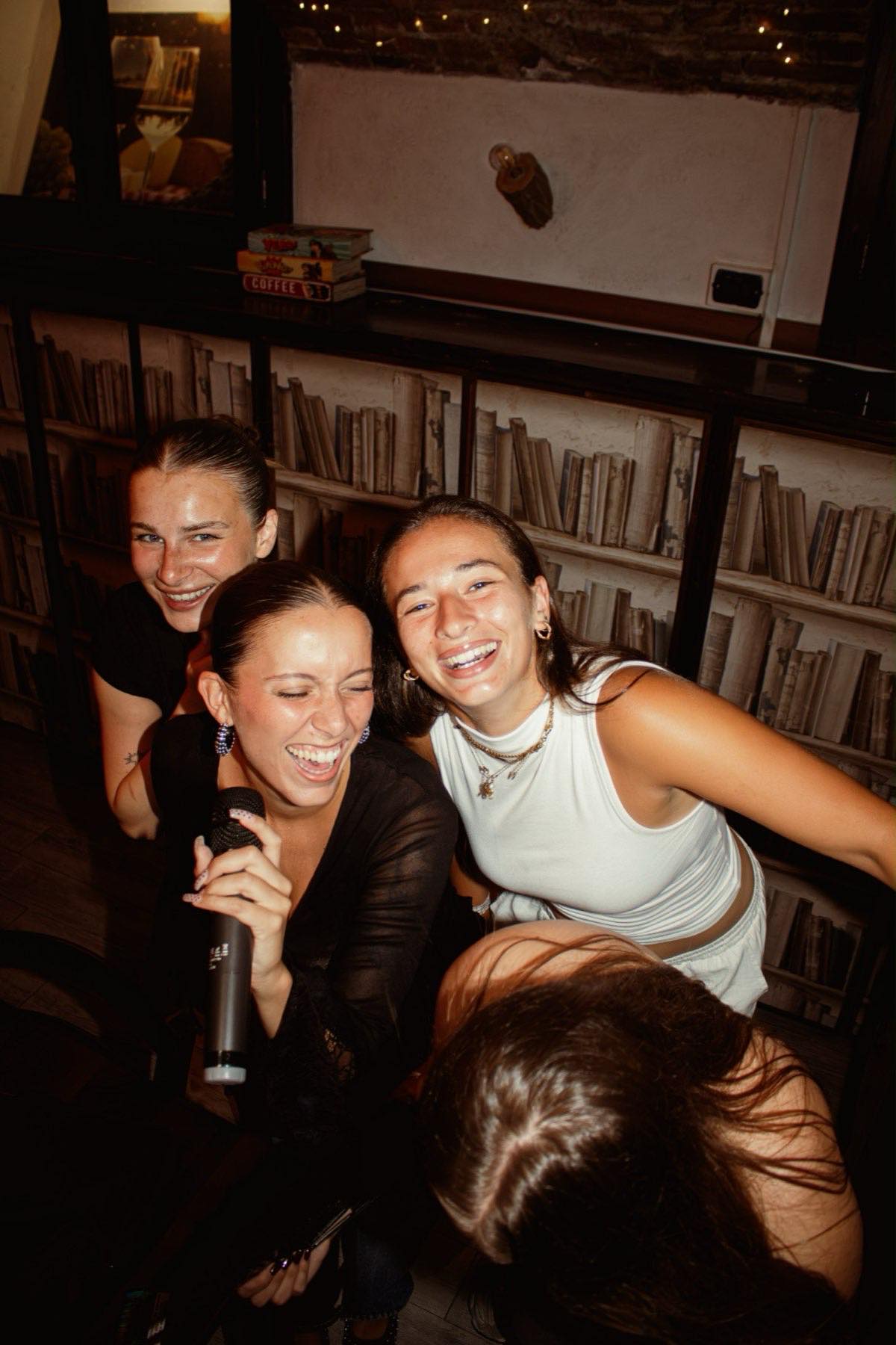 Amiche ridono insieme al microfono davanti a una libreria durante il karaoke