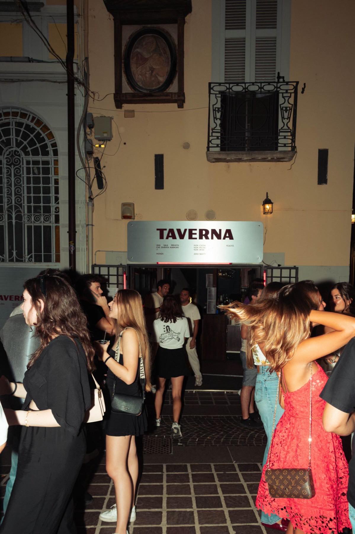 Ingresso di Taverna Felicità con insegna luminosa, ospiti in fila all'entrata